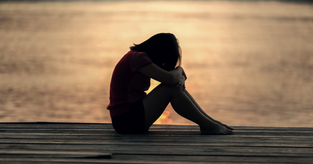 A sad younger lady sitting on a dock with her hands and head folded over her knees